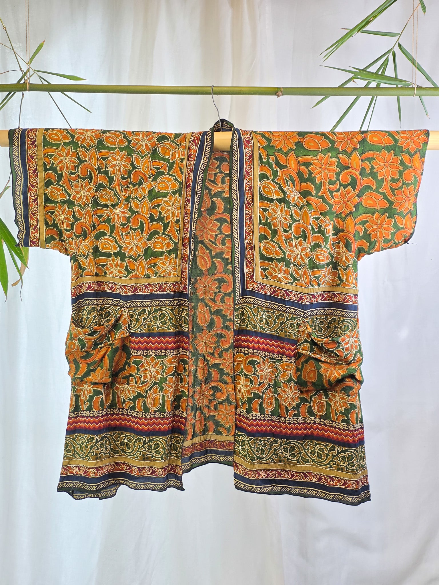 A short-sleeved open-front kimono-style top with a bold orange and green floral print, hanging on a bamboo rod against a white curtain backdrop with decorative bamboo leaves.