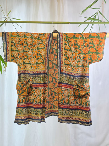 A short-sleeved open-front kimono-style top with a bold orange and green floral print, hanging on a bamboo rod against a white curtain backdrop with decorative bamboo leaves.