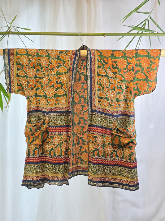 A short-sleeved open-front kimono-style top with a bold orange and green floral print, hanging on a bamboo rod against a white curtain backdrop with decorative bamboo leaves.
