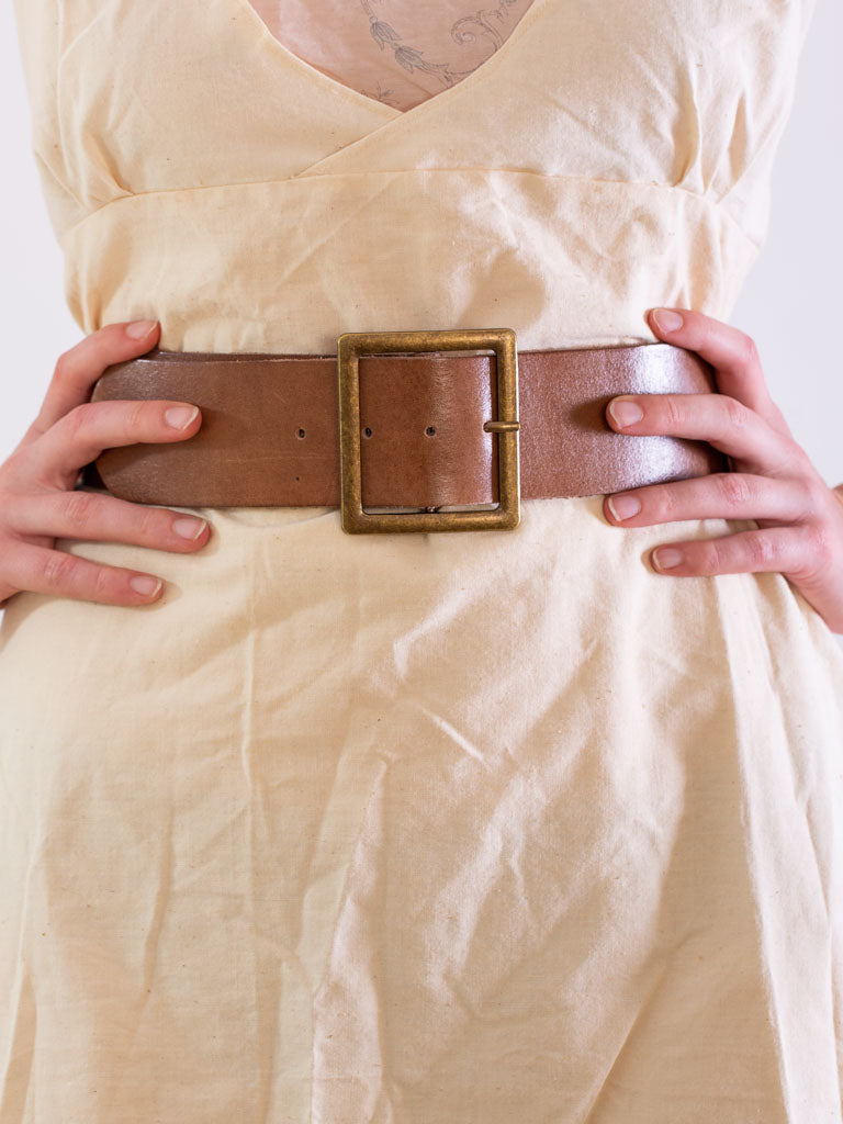 close of models waist wearing a plain tan leather belt with a square brass buckle.