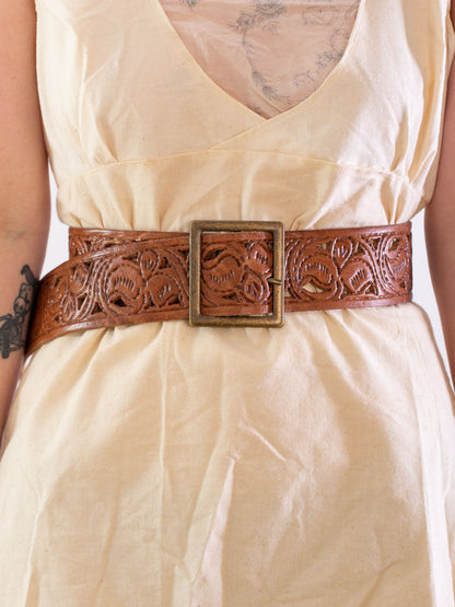 close up of models waist wearing a wide natural coloured leather belt with a square brass buckle.The leather has a scroll design tooled into it.