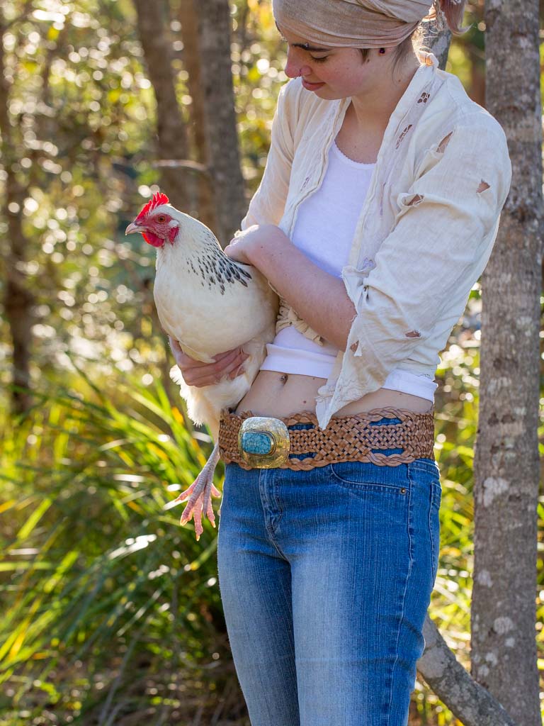 Model holding a chicken wearing a wide woven tan coloured leather belt with an oval buckle engraved an set with a blue chip centre. 